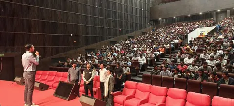 A man with a microphone addresses a large student audience seated in a tiered auditorium with red chairs.