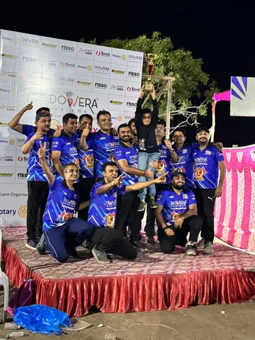Group of men in blue jerseys celebrating on a stage as a child lifts a trophy, sponsor backdrop behind.