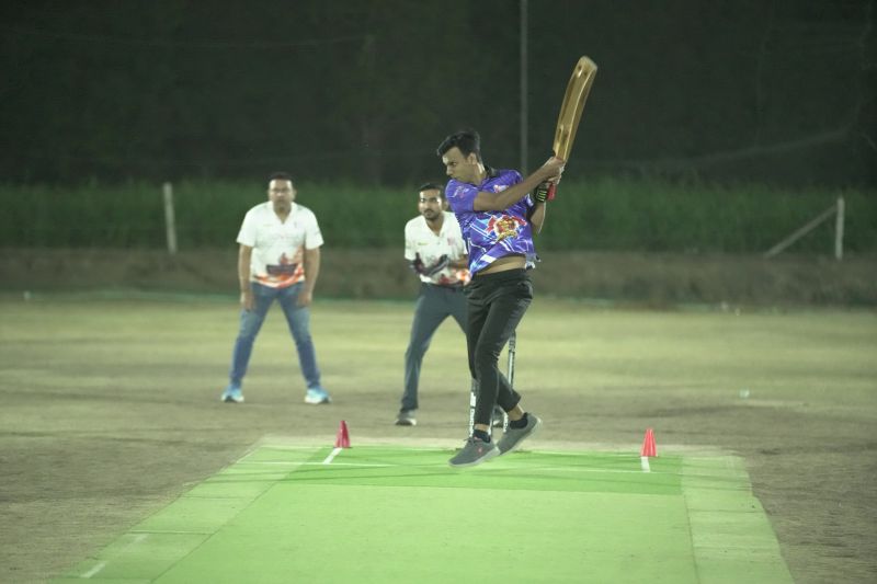 Batsman in blue jersey hitting a shot during a night cricket match on a green mat pitch.