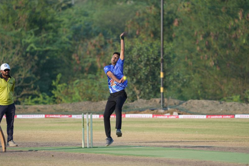 Bowler in blue jersey leaping to deliver the ball beside the stumps at an outdoor cricket ground.