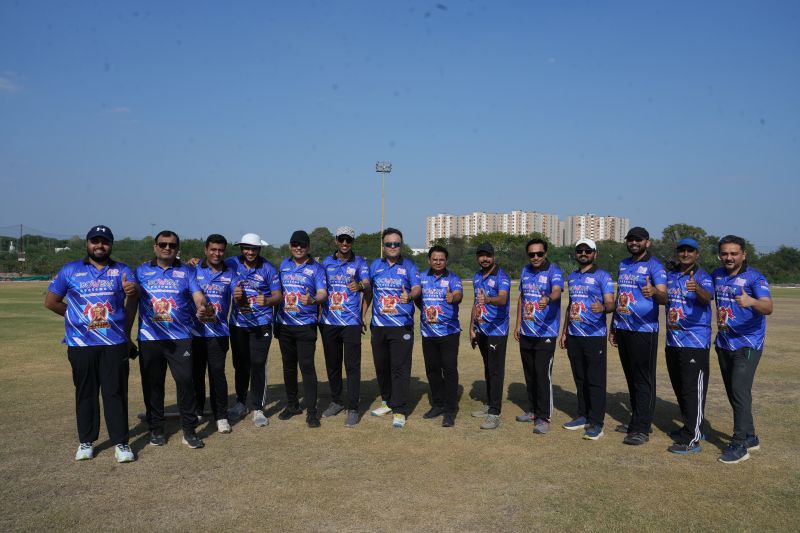 Team of men in matching blue jerseys posing with thumbs up on a grass cricket field.
