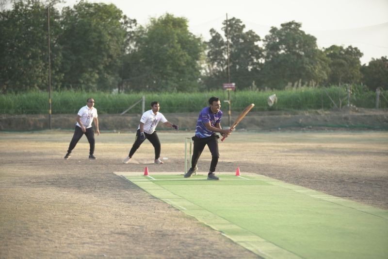 Batsman swinging at a ball on a green mat pitch as gloved fielders wait behind during a daytime match.
