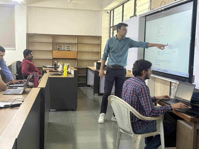 A man points at a large display while students work on laptops around him in a classroom.