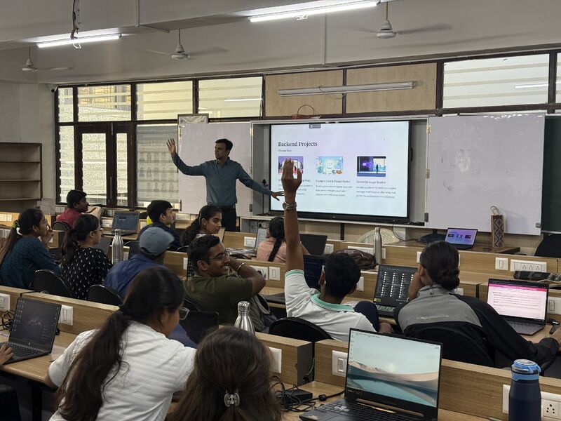 A presenter gestures at a screen listing backend project options while a student raises a hand.