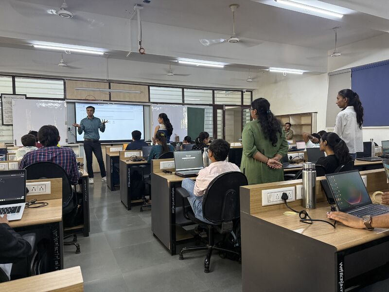 Wide view of a classroom session with a presenter at the front and students on laptops at long desks.