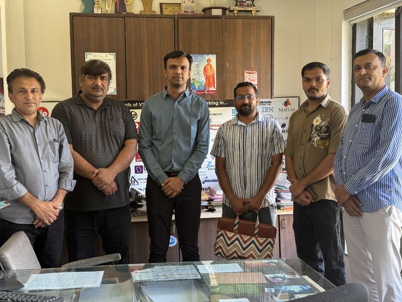 Six men pose together in an office in front of a cabinet and notice board.