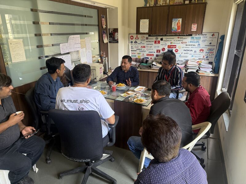 Men talk over snacks around a glass table in an office, one wearing a 'we_must_ship' t-shirt.