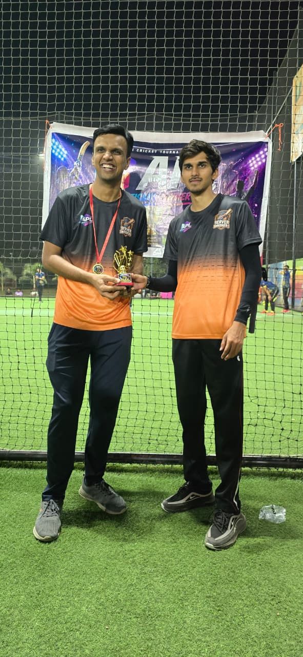 Two smiling men in black-and-orange jerseys hold a small trophy, one wearing a medal, at a night cricket venue.