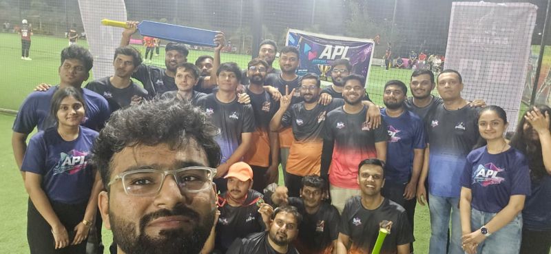 A man with glasses in the foreground takes a selfie with a large group of players, one holding a blue bat.