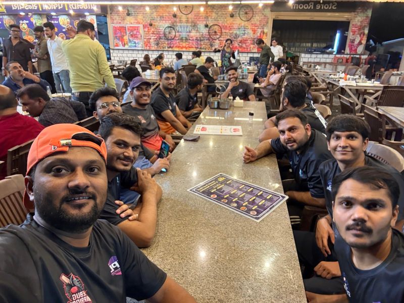 A man in an orange cap takes a selfie with teammates seated around a long table at a restaurant.