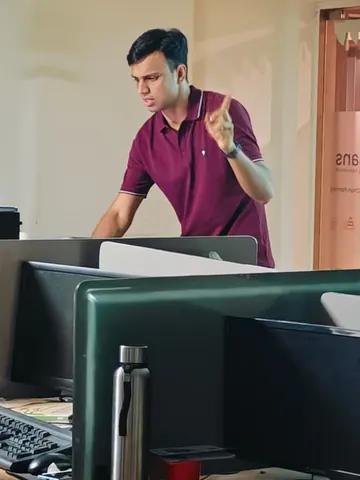 Gaurav gestures while speaking among office desks and monitors during a learning session.