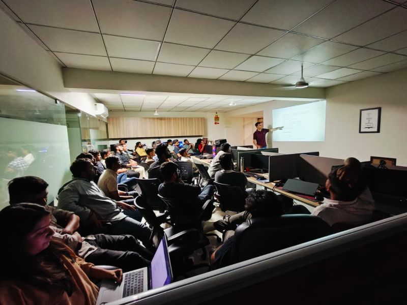 An audience sits at desks as a presenter points to a projected slide in a dim training room.