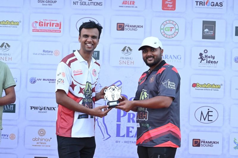 Two men in cricket jerseys share a smile while holding a small trophy in front of a sponsor backdrop.