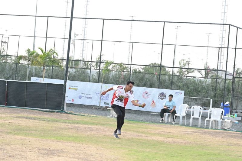A bowler in a red and white jersey delivers on a grass cricket field ringed by nets and floodlight towers.