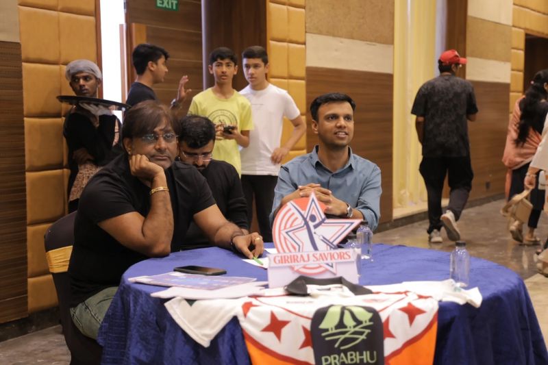 Men seated at a round table with a Giriraj Saviors plaque and cricket jerseys at an indoor gathering.