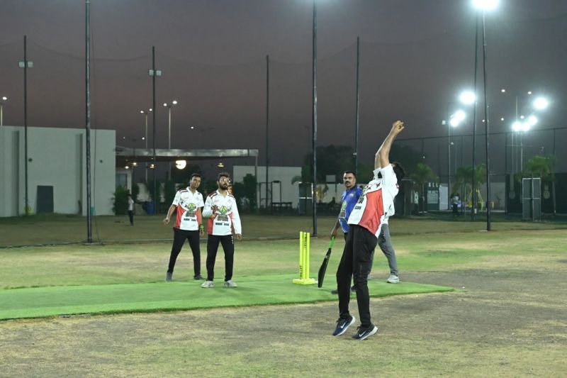 A bowler leaps through his delivery under floodlights at night as a batsman waits at yellow stumps.