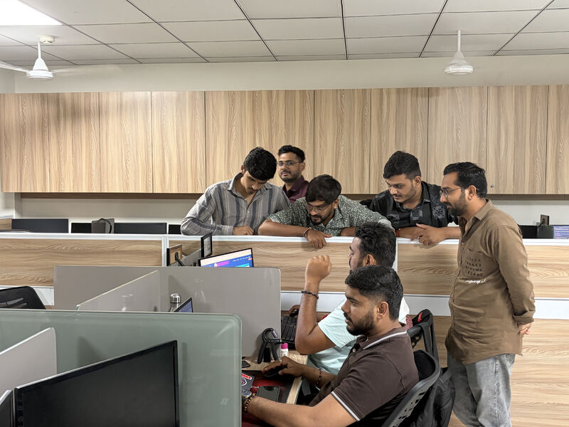 Seven men in an office gather around a desk, one seated and typing while the others look at a monitor.