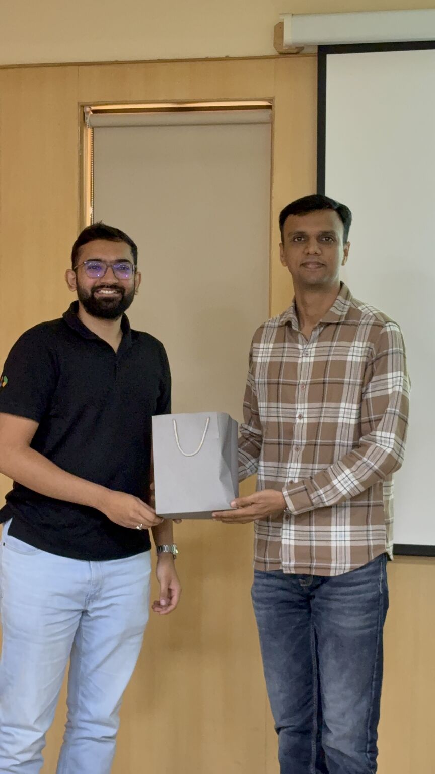 Two men pose indoors holding a grey gift bag together.
