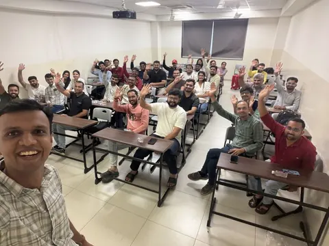 A smiling man takes a classroom selfie with attendees raising their hands at an AI meetup.
